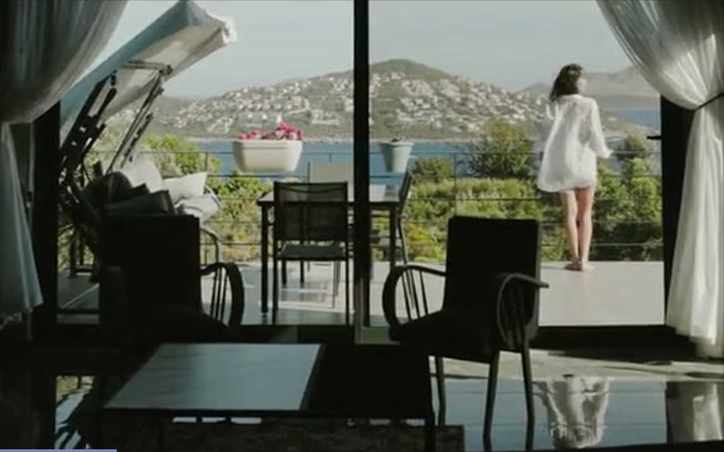A hotel room's floor-to-ceiling window with a woman standing on the terrace outside, hills and water beyond