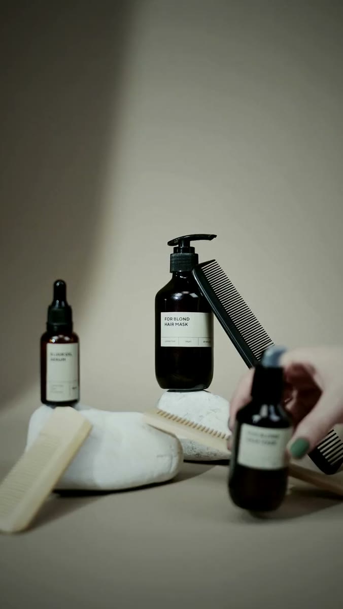 Two amber cosmetic bottles and four combs arranged on a beige backdrop, the taller bottle labeled FOR BLOND HAIR MASK and raised on a white stone, a black comb leaning against it, and a hand with mint-green nail polish lifting a third bottle into the foreground